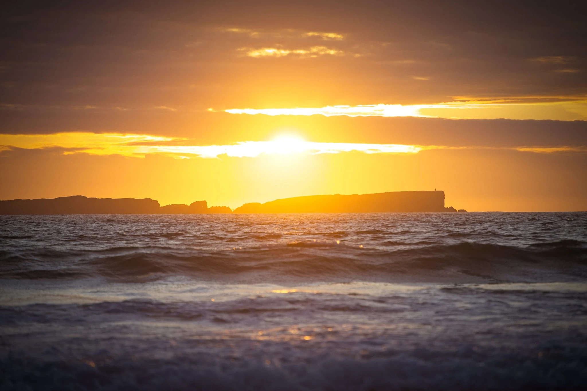 Shot from near sea level, waves roll past the camera toward the silhouetted Berlengas Islands as the sun breaks through thick cloud in a last burst of gold and orange.