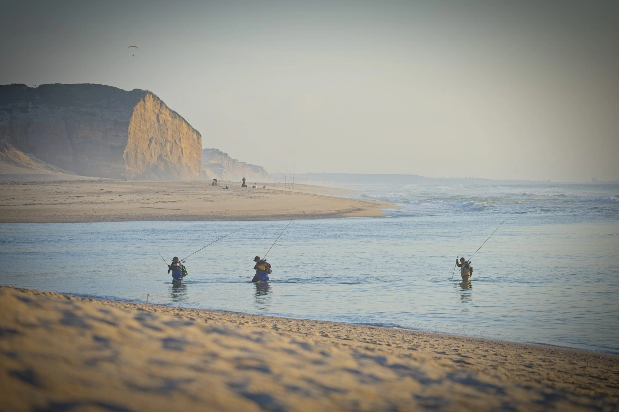 Three fishermen stand waist-deep in the tidal channel, rods arcing toward the sea, sandy cliffs behind and a paraglider drifting high overhead — a layered scene at the river mouth.