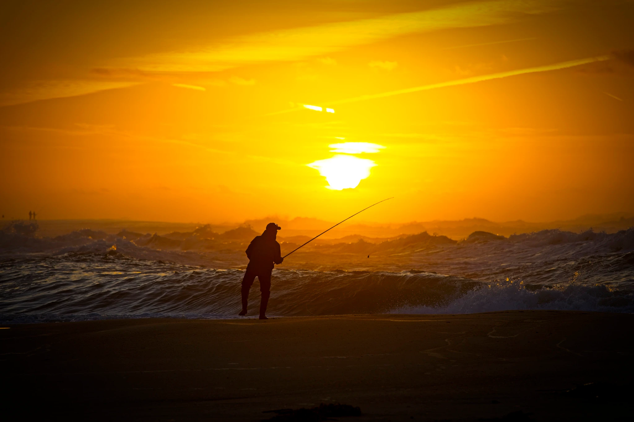 A lone fisherman holds their ground at the shoreline, rod bent and extended toward the sea, as a dramatic orange sunset blazes behind them and waves churn past their feet.