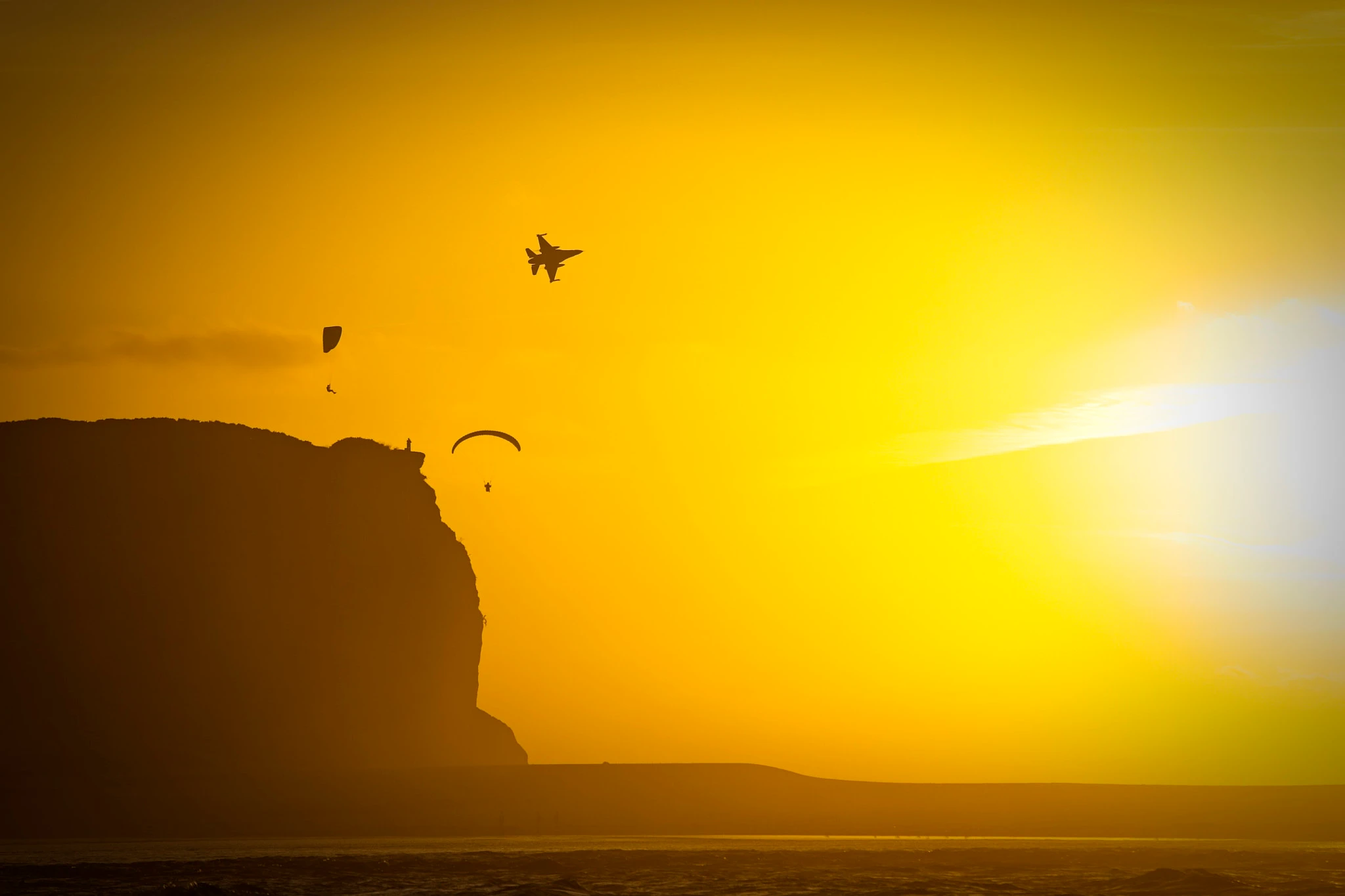 Two paragliders launch off the edge of a cliff into a golden haze as a fighter jet crosses directly above — slow and fast sharing the same blazing sky by pure coincidence.