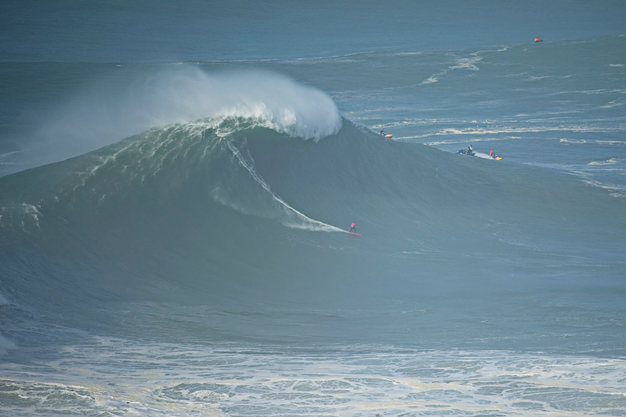 A surfer carves down the face of a towering Nazaré wave, the wall of grey-green water rearing above them at terrifying scale, a jet ski standing by on the far side.