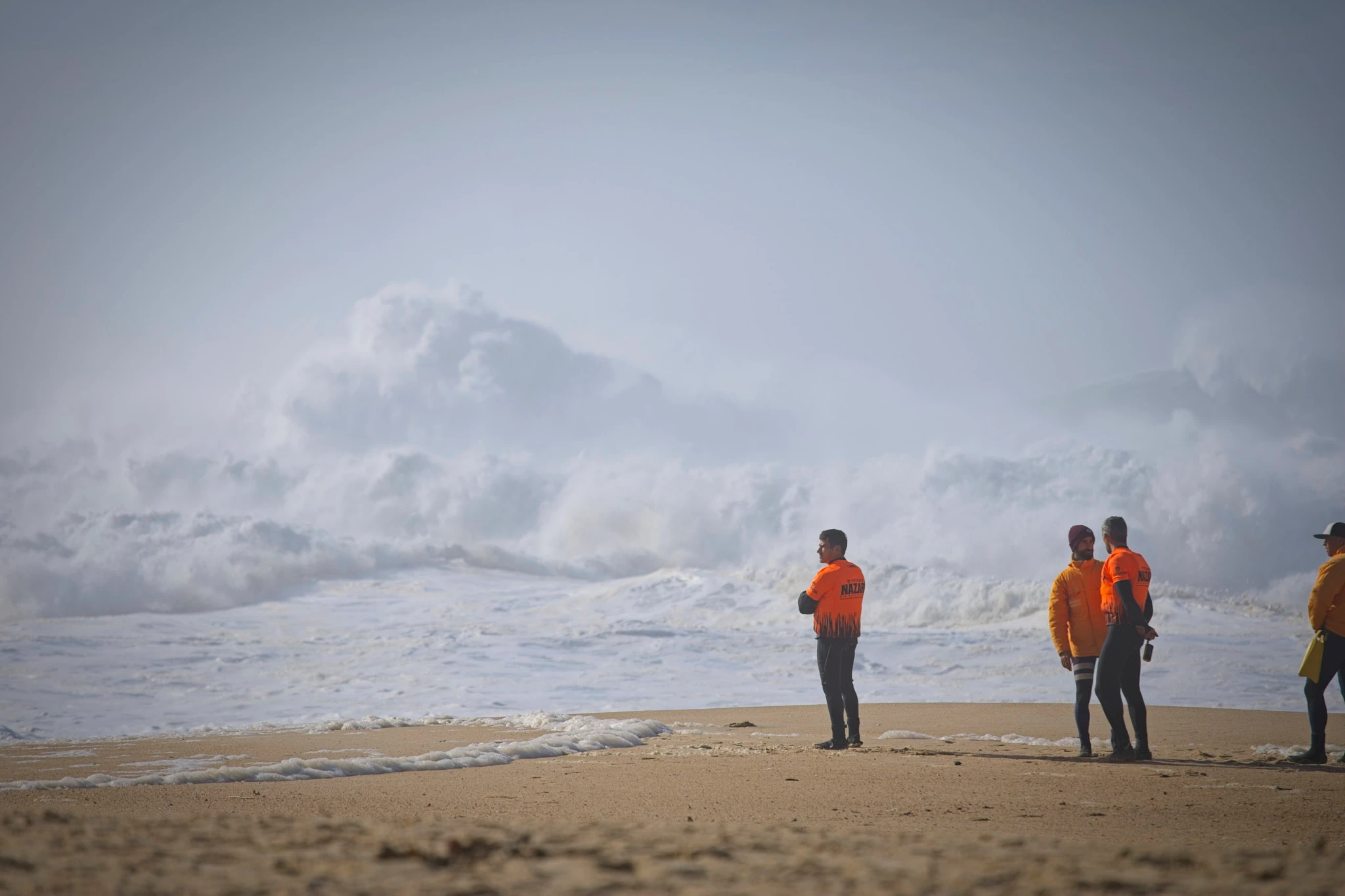 Water safety crew in orange Nazaré vests stand on the beach watching enormous waves crash on the shoreline, mist and spray filling the air behind them in a wall of white chaos.
